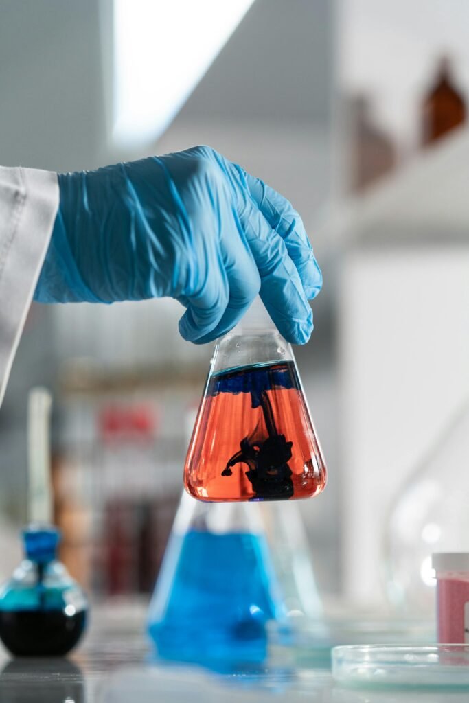 oil-1 Close-up of hand in glove holding a flask with colored liquid in a laboratory setting.